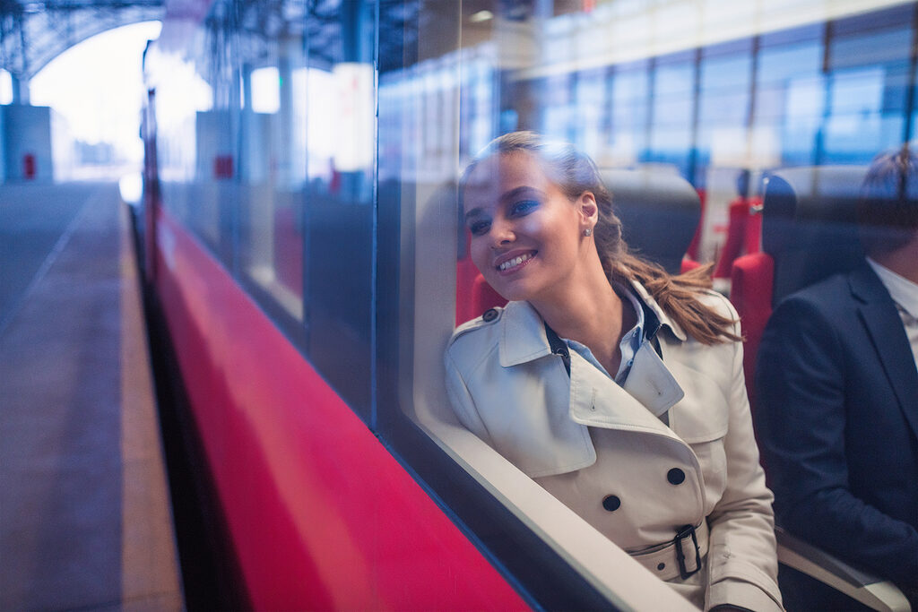 Außenansicht einer lächelnden Person, die am Fenster eines Zugwagons lehnt und hinausblickt